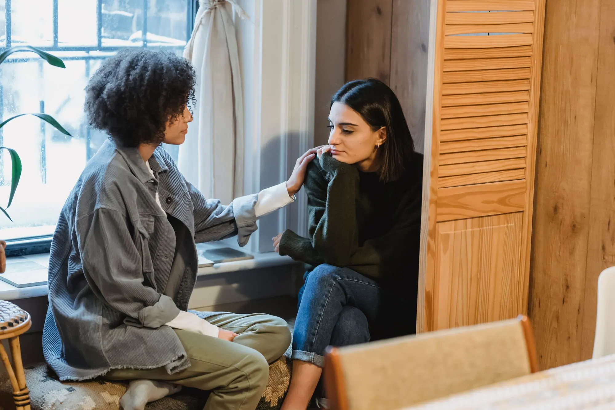 Woman resting a reassuring hand on a friend’s shoulder as they talk quietly by a window